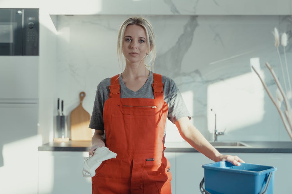 A female cleaner with blonde hair, dressed in a grey t-shirt and red overalls, standing in a modern kitchen during daytime. She holds a white cleaning cloth in her right hand and rests her left hand o